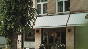 Un bâtiment à la façade en briques, doté de grandes fenêtres et d’une entrée abritée. On y trouve des espaces aménagés pour s’asseoir à l’extérieur et des plantes en pot.