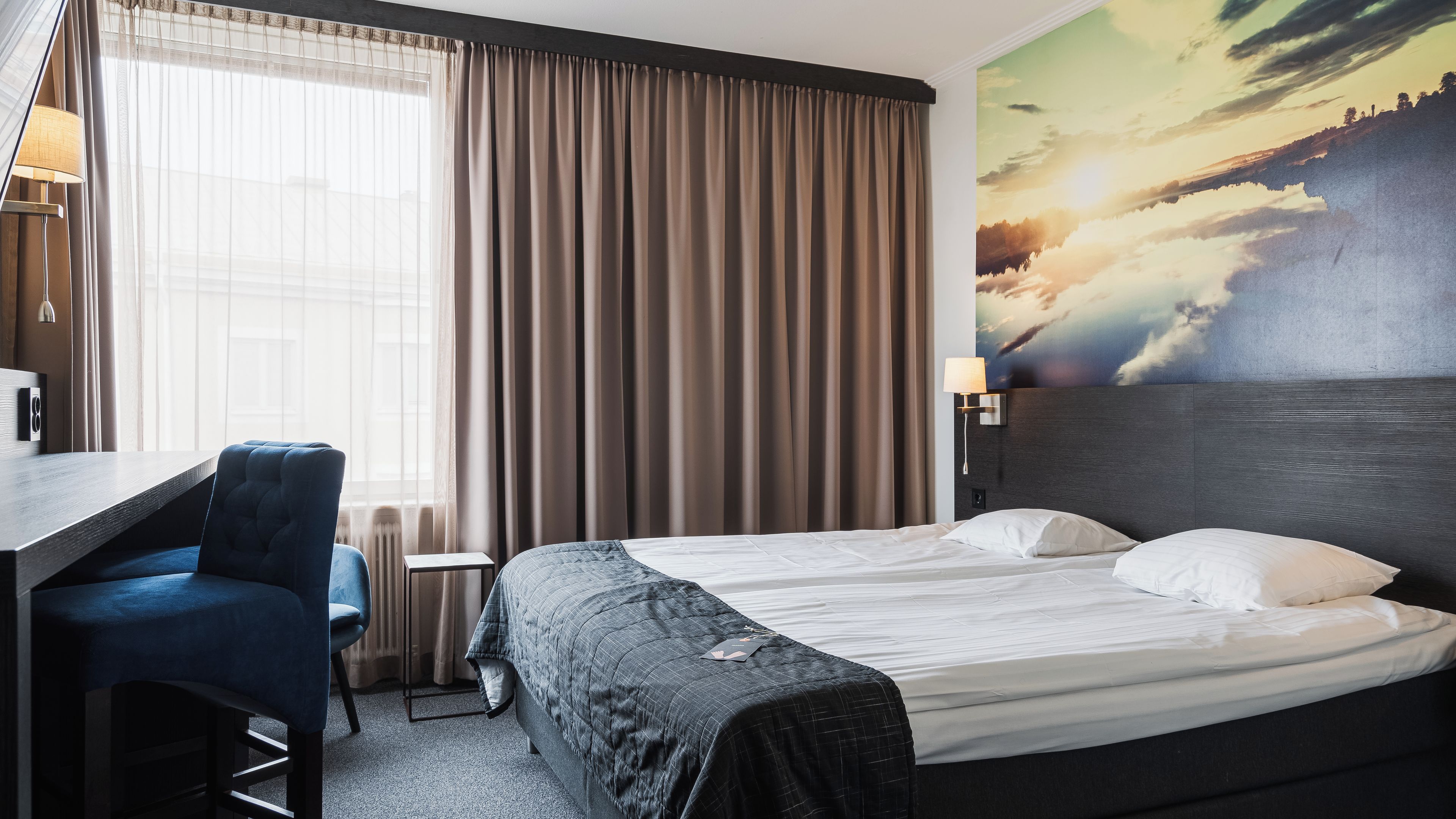 Habitación de hotel con cama, escritorio, silla, lámpara y un cuadro de un atardecer.