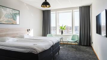 A hotel room with a bed, a chair, a table, a large window, and a painting on the wall.