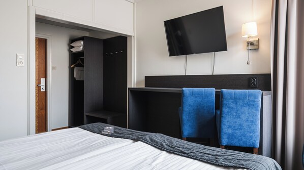 Quarto de hotel com cama, televisão fixada na parede, guarda-roupa e casa de banho visível por uma porta aberta.