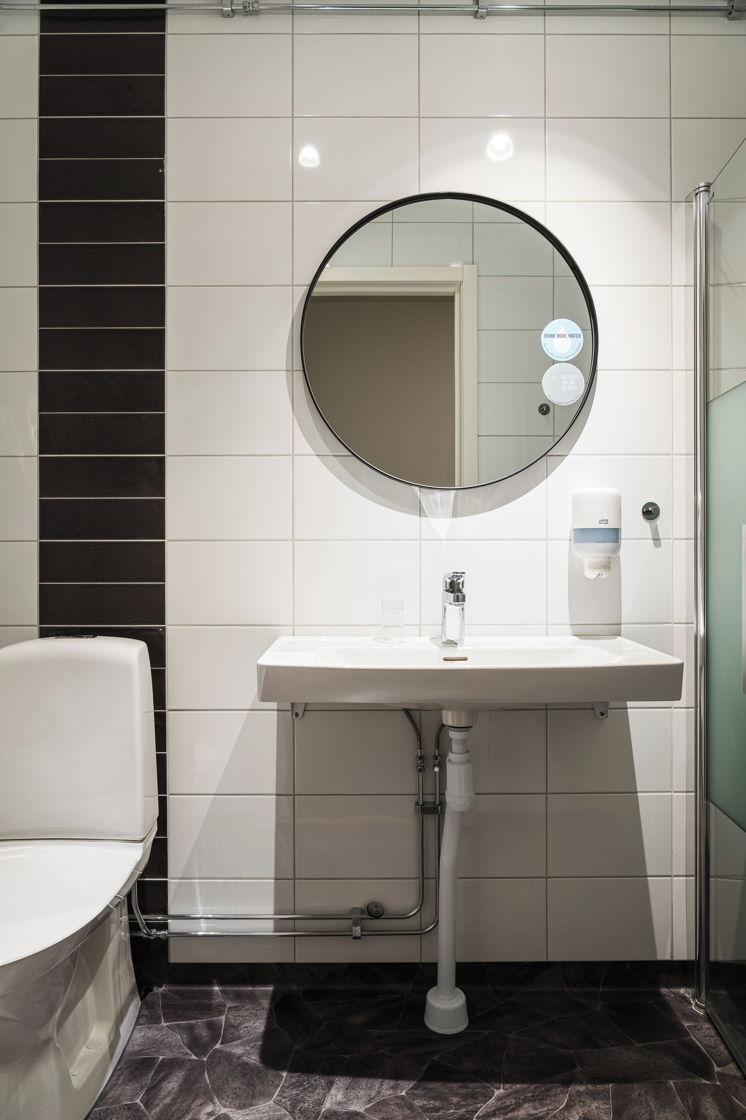 A bathroom with a white sink, a round mirror, a soap dispenser, and a toilet.