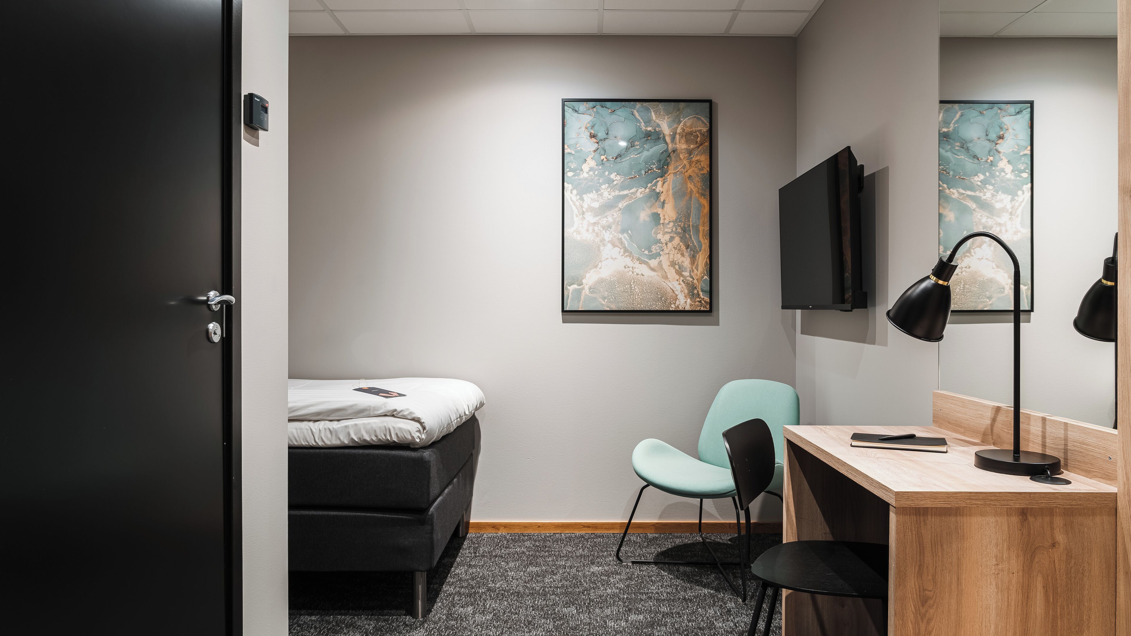 A hotel room with a bed, a desk, a chair, a TV, and artwork on the walls.