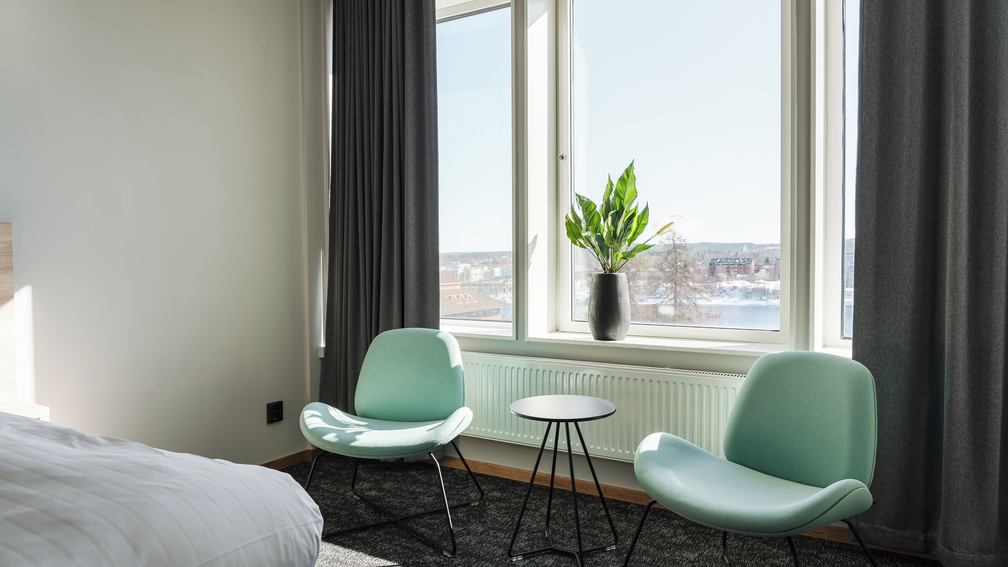 Habitación de hotel con cama, dos sillas, una mesita, una planta en maceta y ventana con vista a la ciudad.
