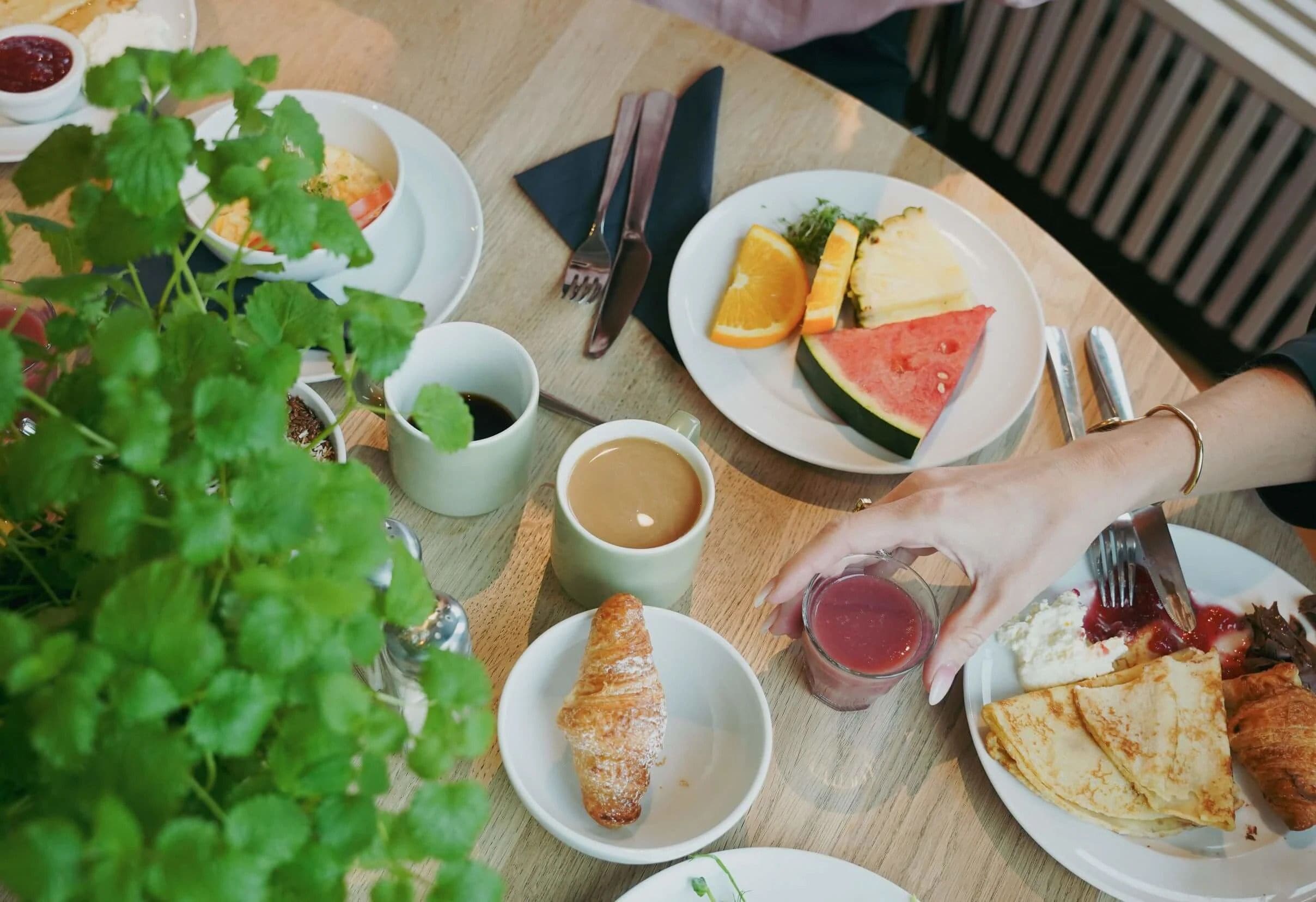 A table with plates of food, including a croissant, a slice of watermelon, and a plate with orange slices and a green salad. There are also cups of coffee and a glass of red juice.
