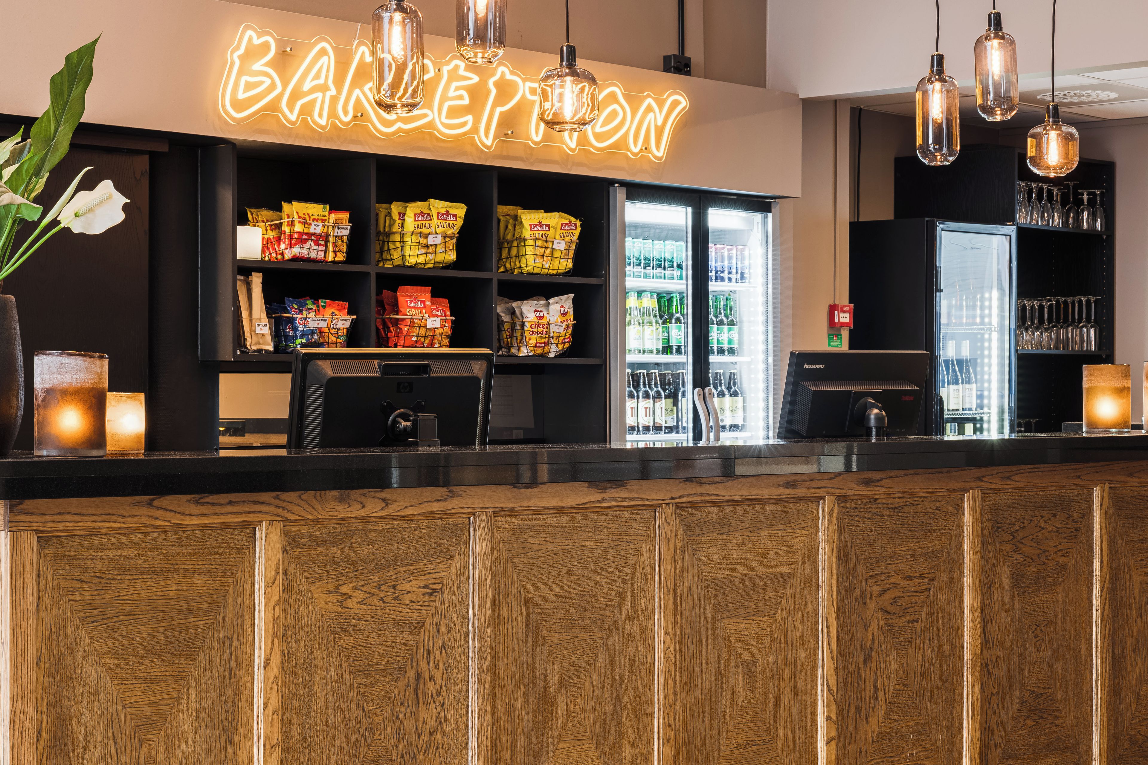 A bar area with a neon sign, shelves stocked with snacks, a refrigerator with beverages, and hanging pendant lights.