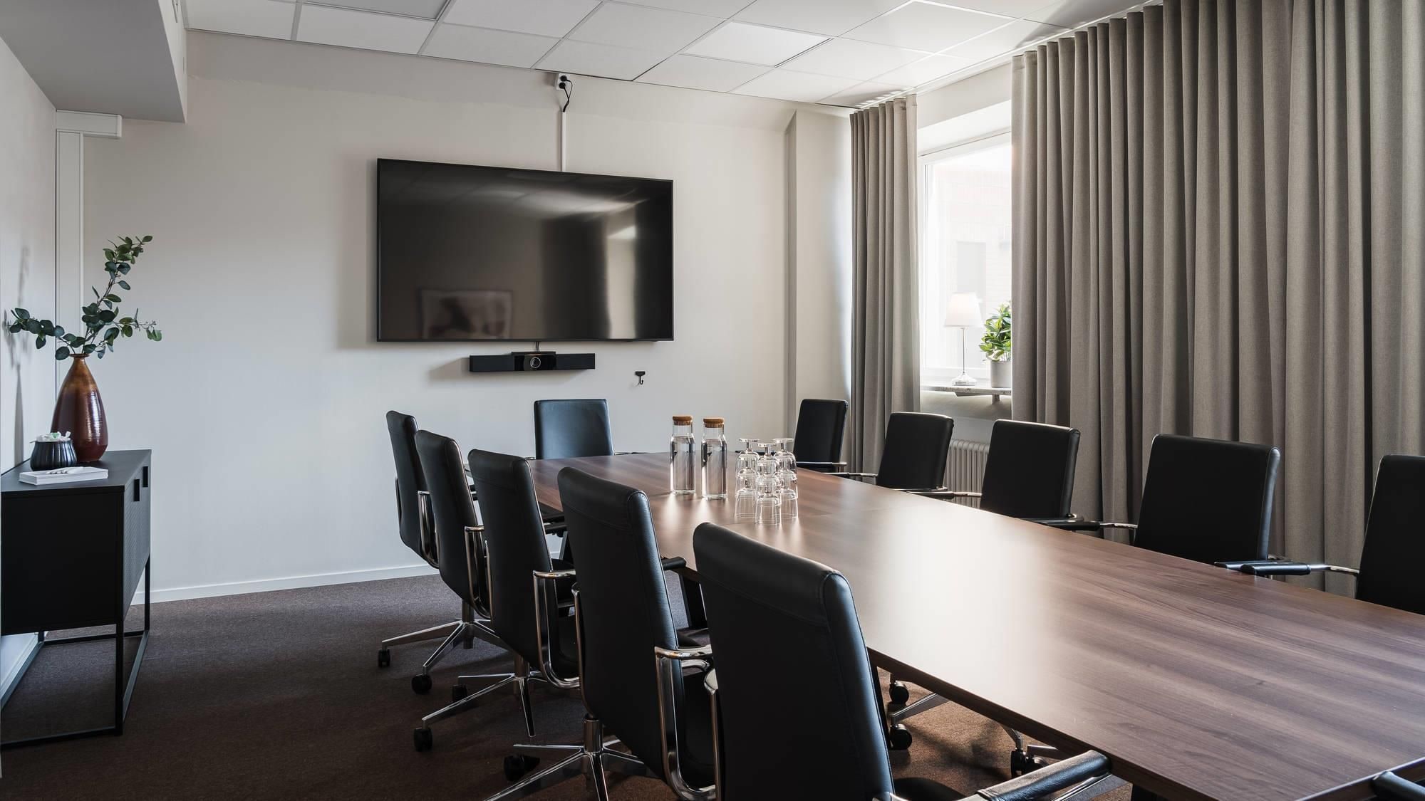 Uma sala de conferências com uma mesa comprida, cadeiras, uma TV de tela plana e um amplo janelão com cortinas.