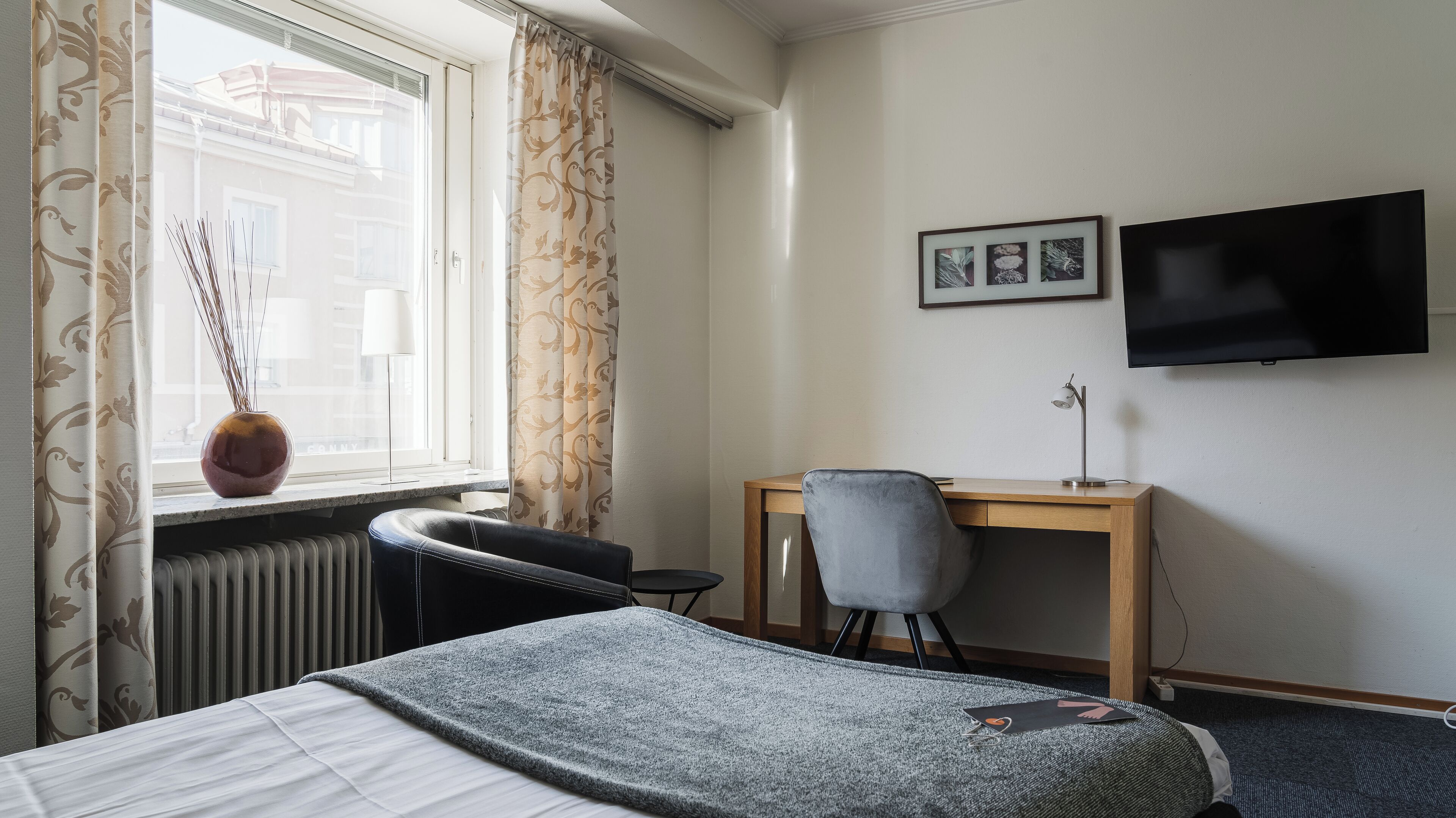 Habitación de hotel con cama, escritorio, silla, radiador, ventana con cortinas, un jarrón con cañas y un televisor de pantalla plana en la pared.