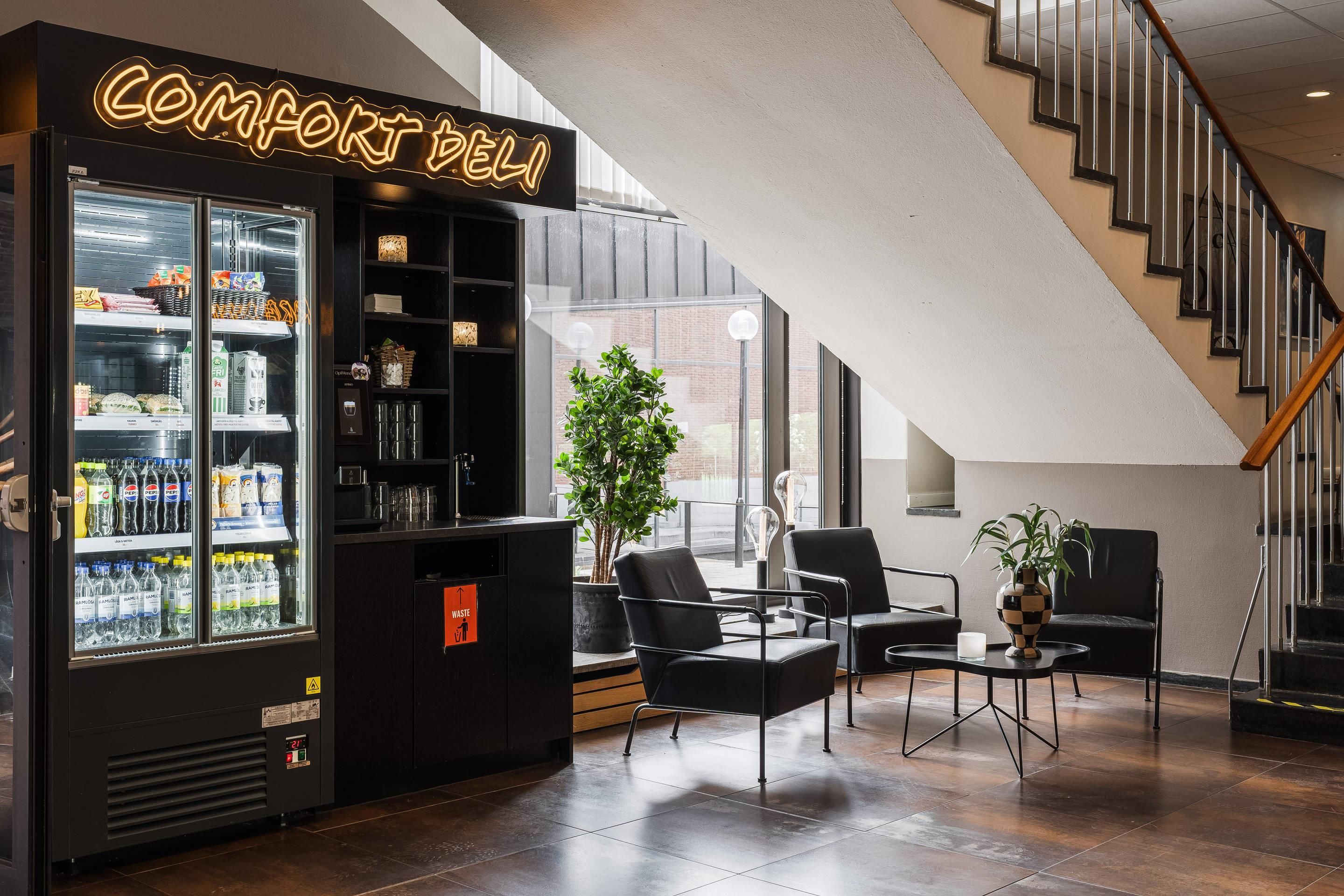 En modern inredning med en försäljningsautomat märkt ”COMFORT DELI”, en sittgrupp med stolar och ett litet bord samt en trappa.