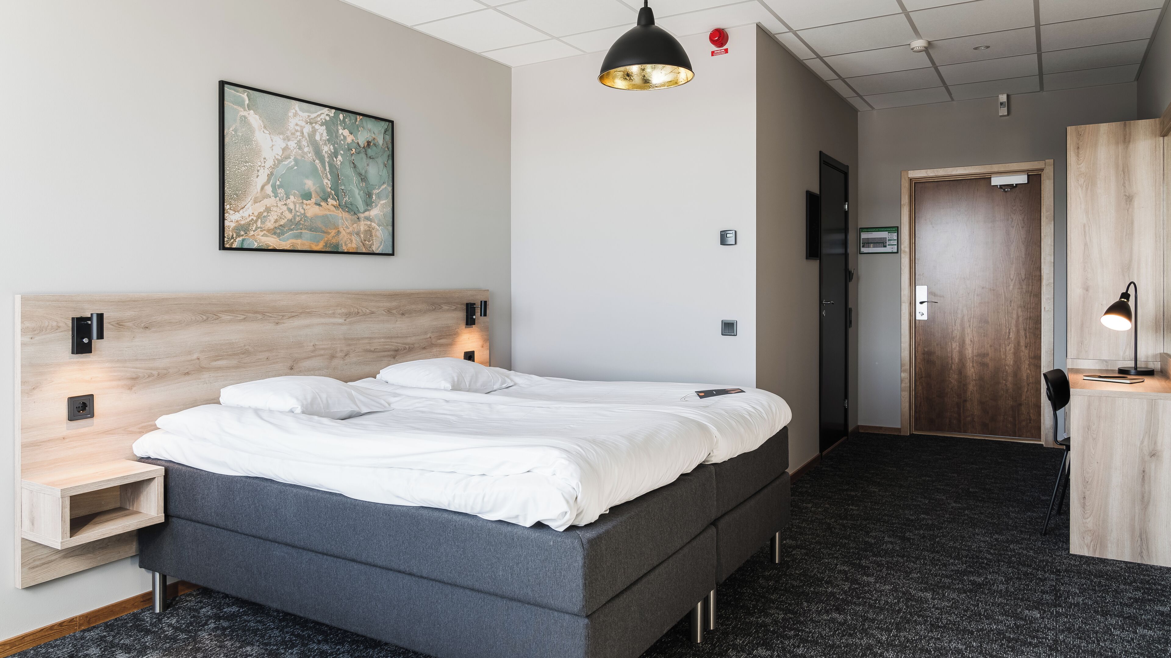 A modern hotel room with a large bed, a wooden headboard, a desk with a chair, and a painting on the wall.