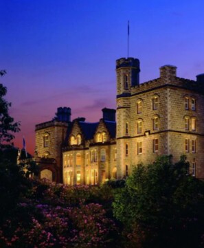 A large, historic stone building with multiple towers and arched windows, surrounded by lush greenery and flowering plants. - Inverlochy Castle (Fort William)