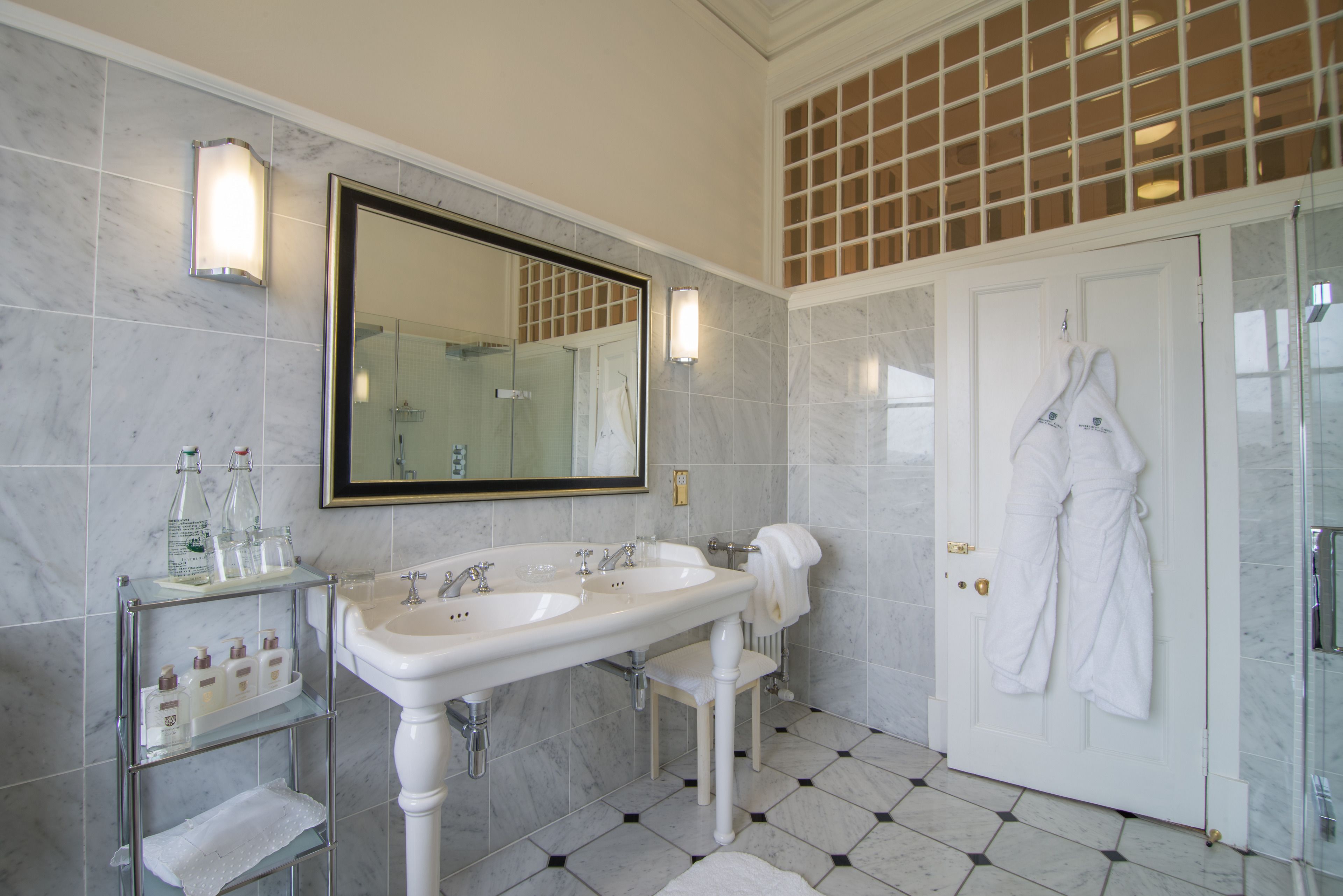 A bathroom with a large mirror, two sinks, a glass shower enclosure, and a white door with towels and robes hanging.
