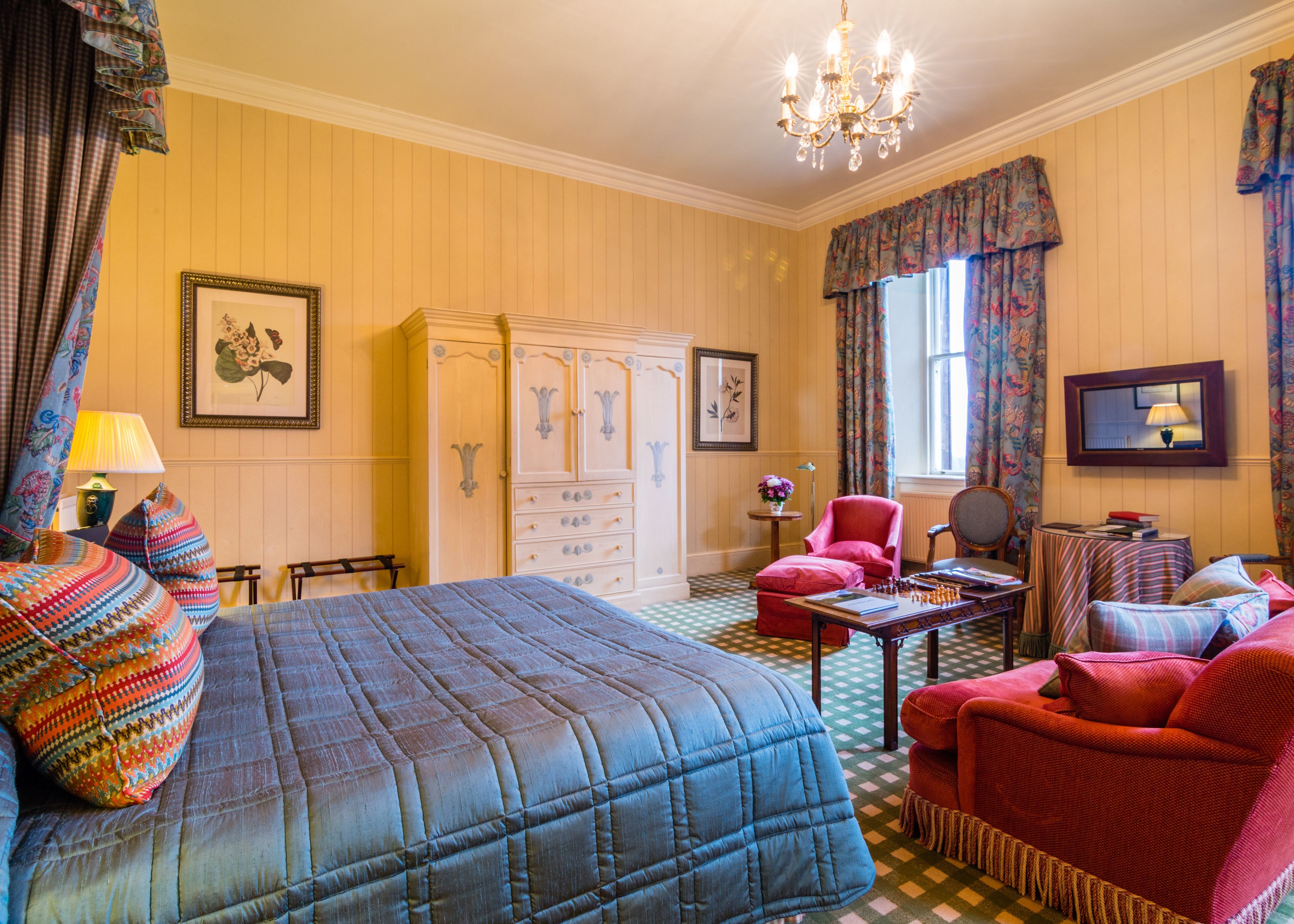 Una habitación con una cama grande, un sillón rojo, una mesa de madera con ajedrez y una araña.