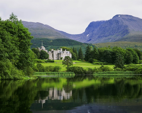 A castle with a lake in the foreground, surrounded by lush greenery and mountains in the background. - Inverlochy Castle (Fort William)