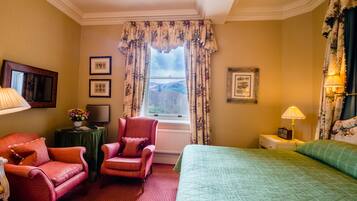 A hotel room with a bed, two chairs, a table with a lamp, a TV, and framed pictures on the wall.