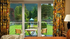 A bedroom with a bed, a table, and two chairs, a balcony with a view of a garden and a gazebo, and curtains with a fruit pattern. - Inverlochy Castle (Fort William)