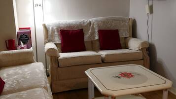 A living room with a beige sofa, a white coffee table with a floral design, a red cushion, and a flat-screen TV mounted on the wall.