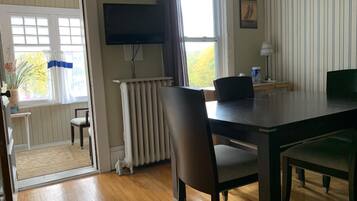 A room with a wooden floor, a dining table with chairs, a radiator, a television mounted on the wall, and a window with curtains.