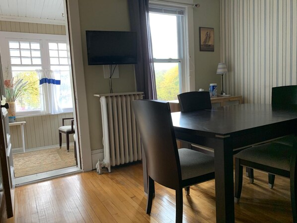 A room with a wooden floor, a dining table with chairs, a radiator, a television mounted on the wall, and a window with curtains.