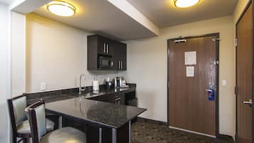 A compact kitchen with a granite countertop, two barstools, a microwave, and a door with a bulletin board.
