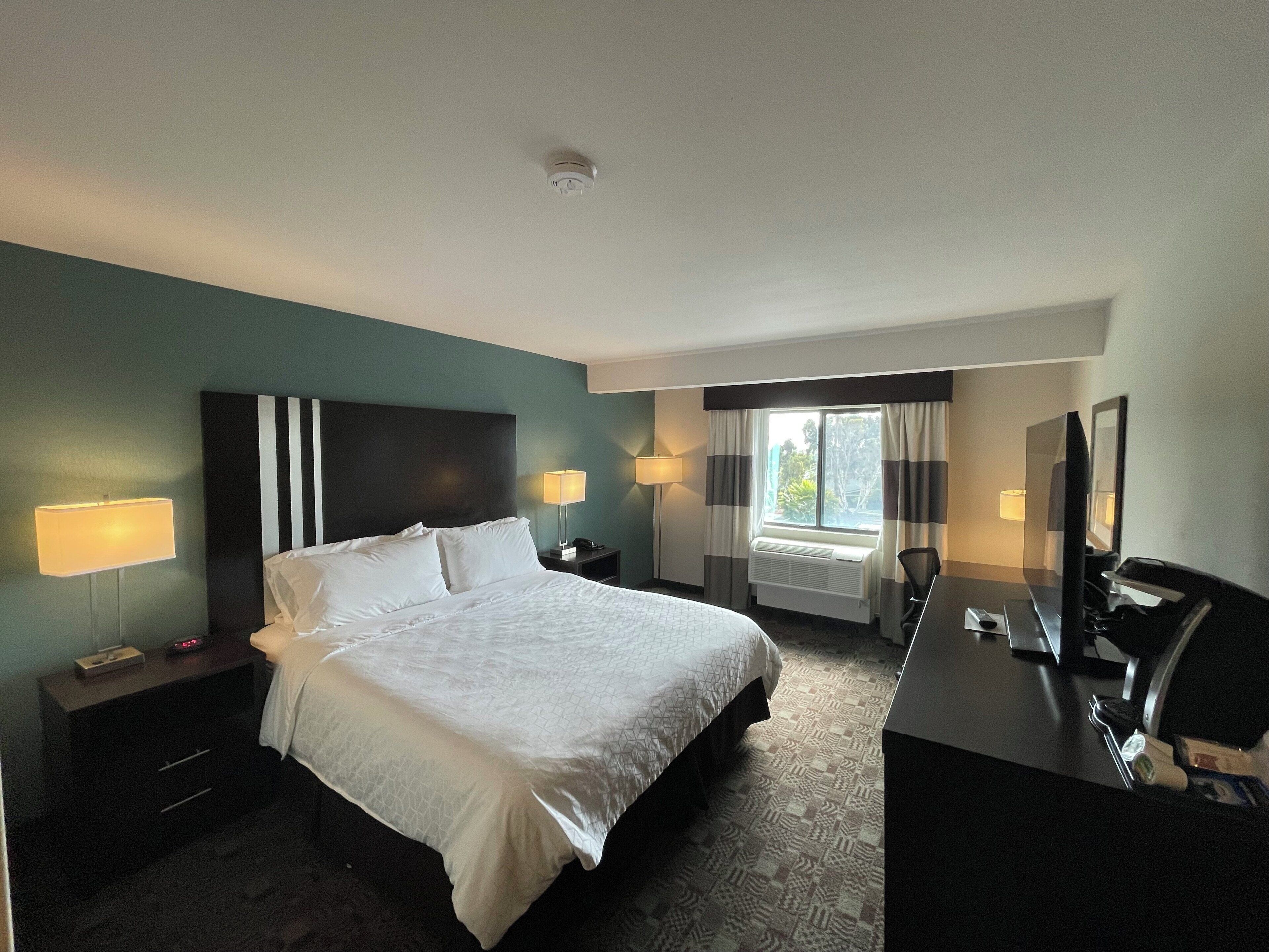 A hotel room with a bed, bedside lamps, a desk with a computer, and a window with curtains.