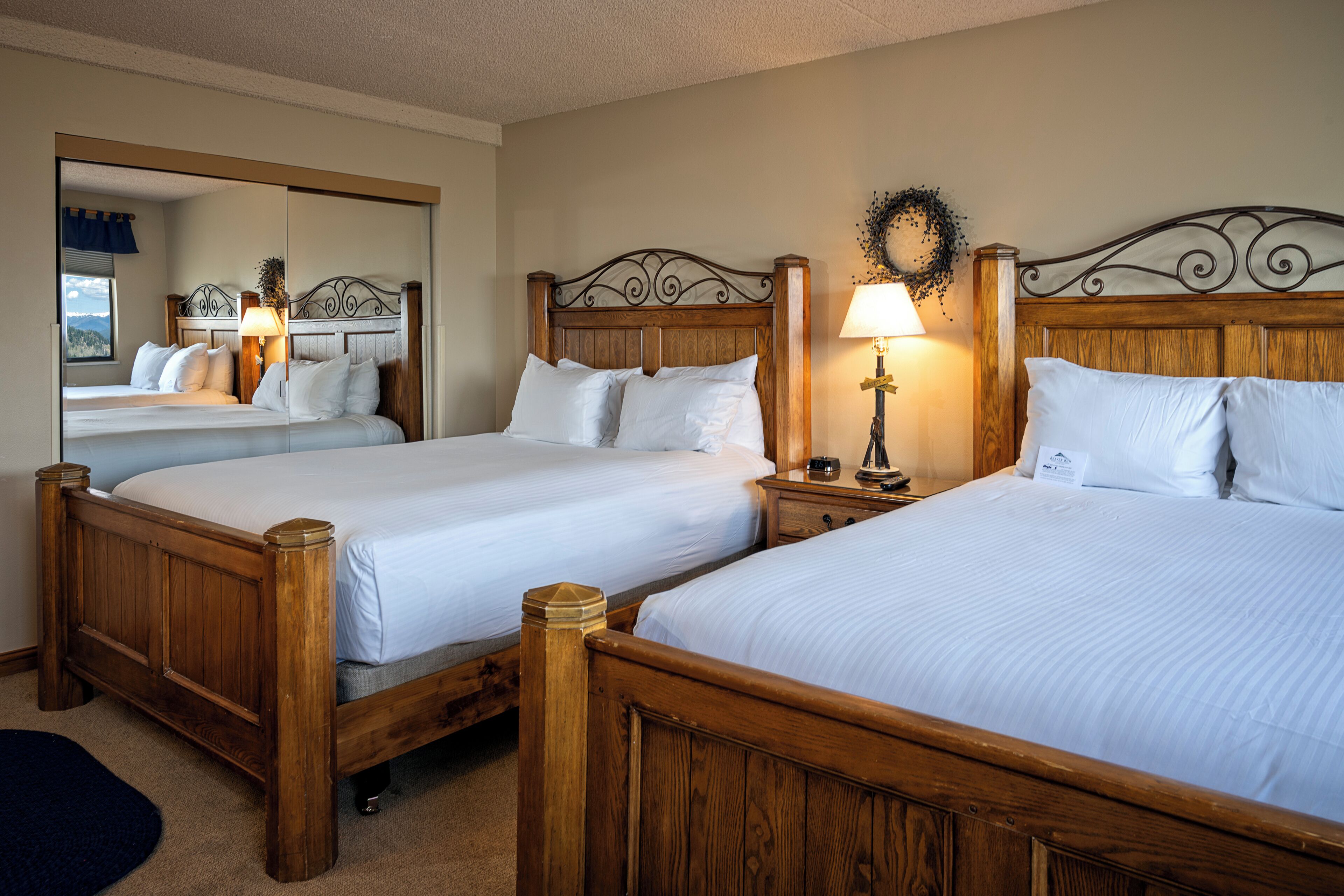 Habitación de hotel con dos camas de madera, una mesita de noche con lámpara y un espejo que refleja la habitación.