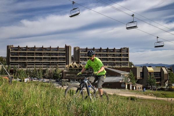 A person riding a mountain bike on a grassy slope with a building and a ski lift in the background.