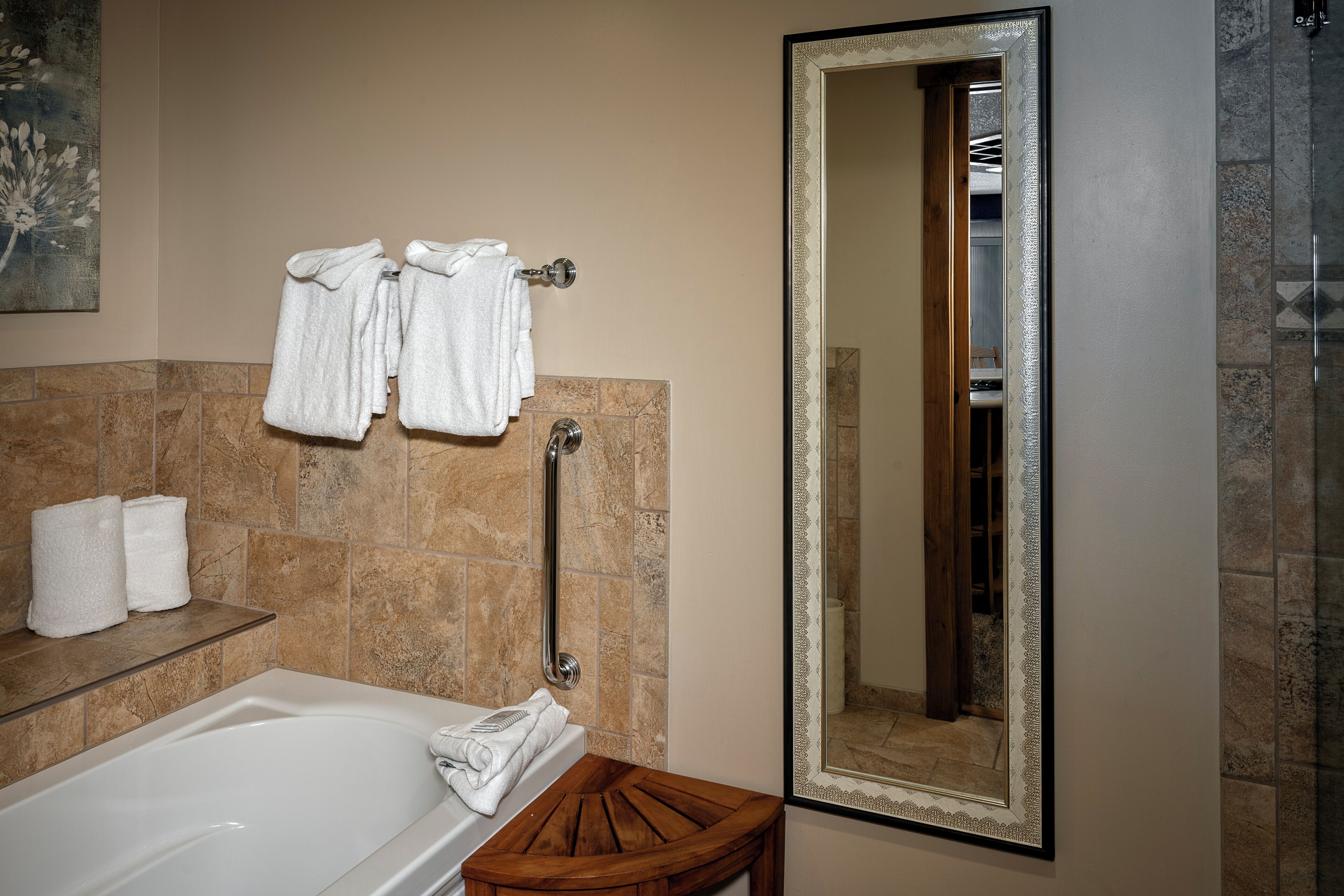 A bathroom with a bathtub, a wooden stool, a towel rack with towels, a handrail, and a mirror with a decorative frame.