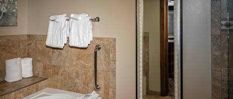 Une salle de bain avec une baignoire, un tabouret en bois, un porte-serviettes avec des serviettes, une barre d’appui et un miroir avec un cadre décoratif.