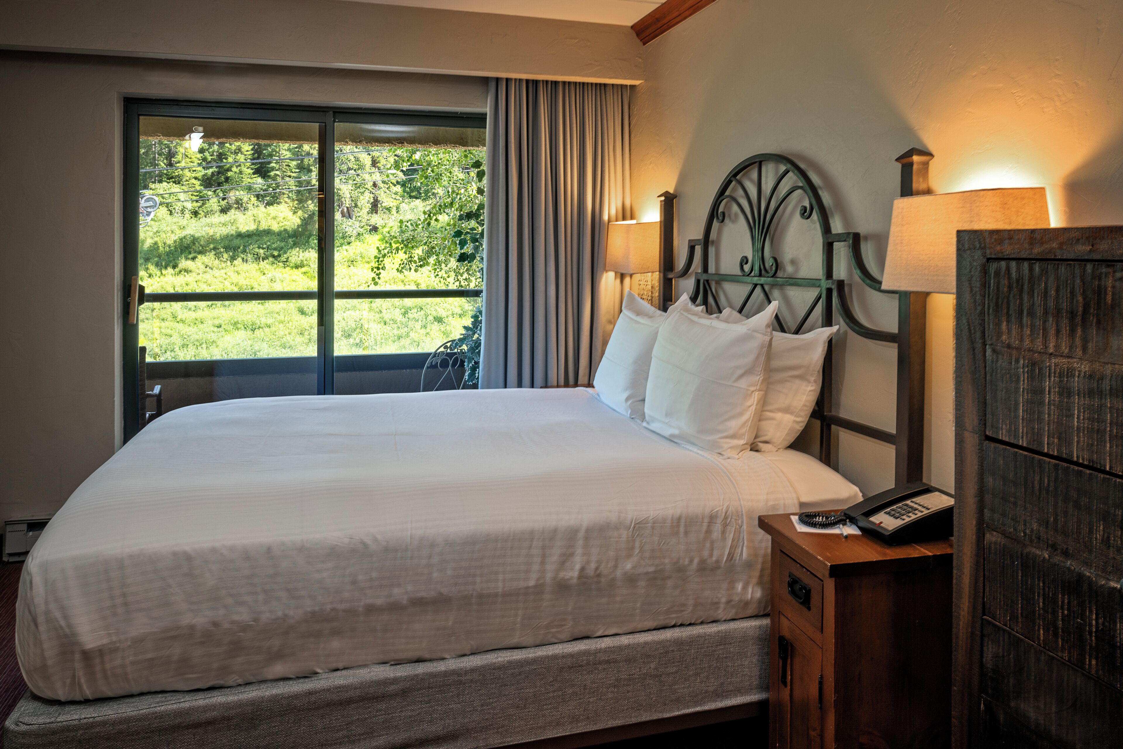 A neatly made bed with white linens, a bedside table with a lamp, and a window with curtains.