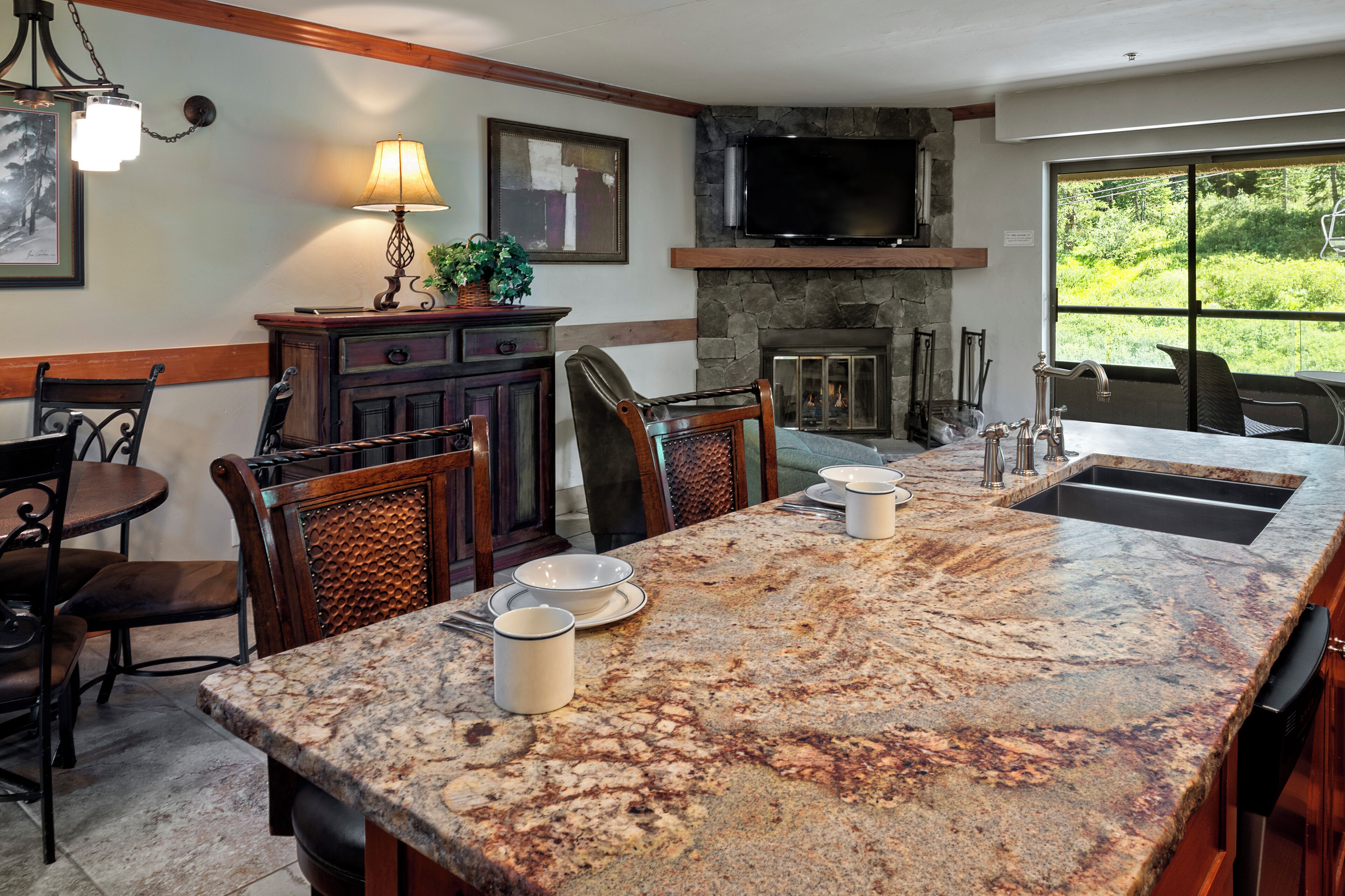 A spacious kitchen with a large granite island, wooden chairs, a dining table, and a stone fireplace.