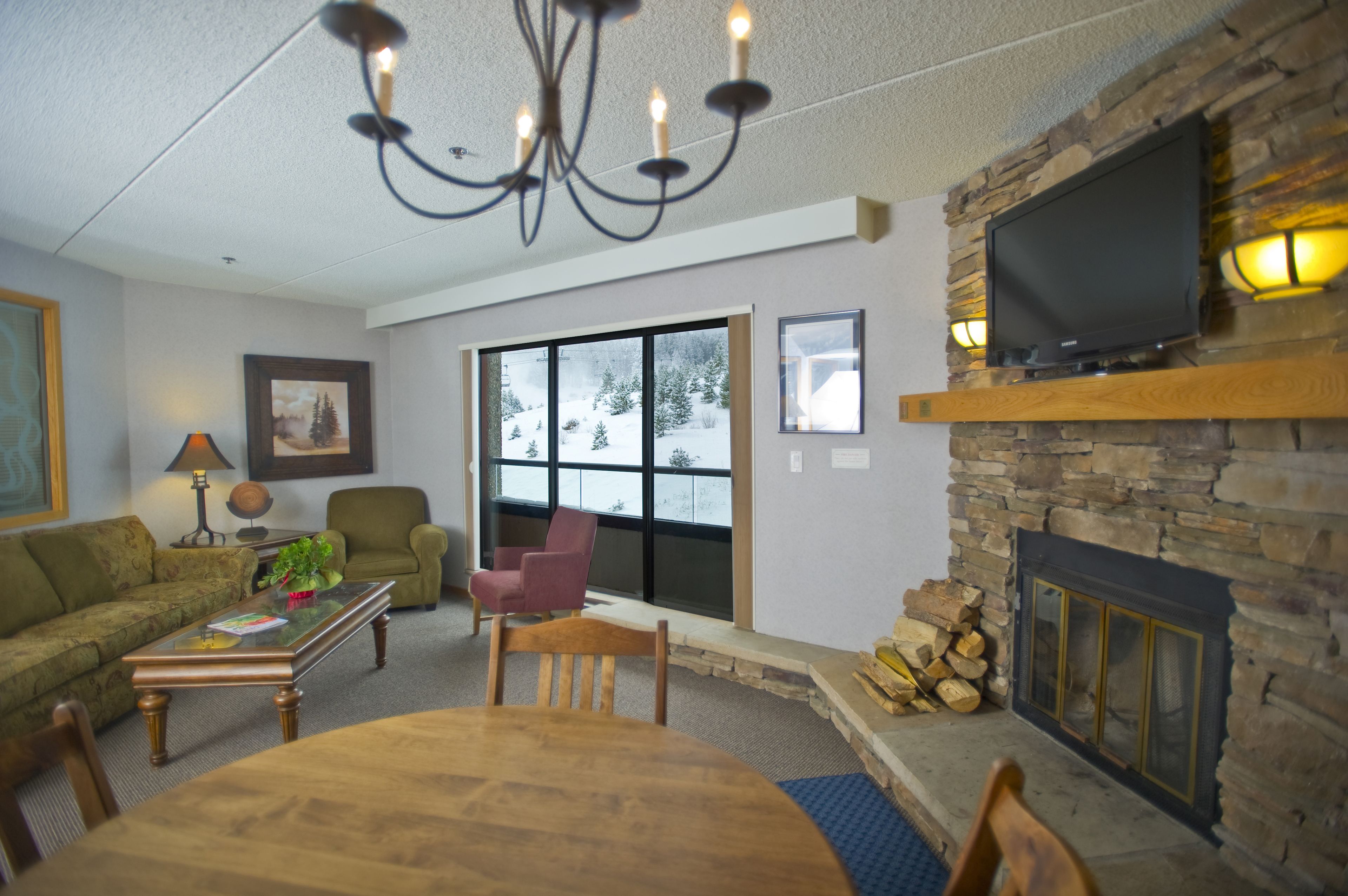 Una sala de estar con chimenea, televisor montado encima, candelabro, sofá, mesa de centro y mesa de comedor con sillas.