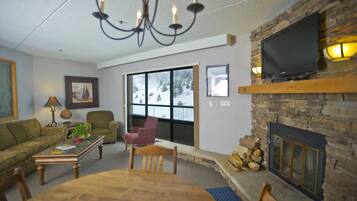 Una sala de estar con chimenea, televisor montado encima, candelabro, sofá, mesa de centro y mesa de comedor con sillas.