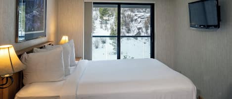 Habitación de hotel con una cama grande, un televisor montado en la pared, una ventana con vistas a árboles nevados y un cuadro en la pared.