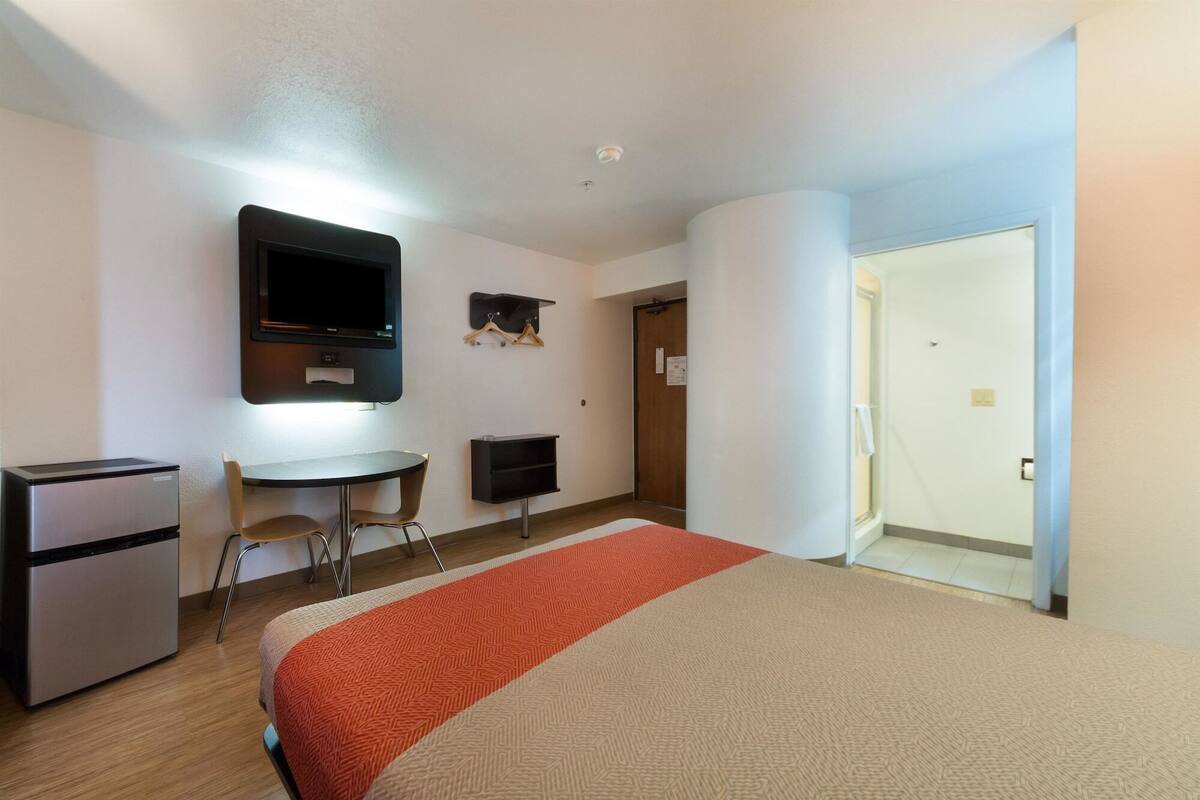 A hotel room with a bed, a desk, a chair, a refrigerator, and a TV.