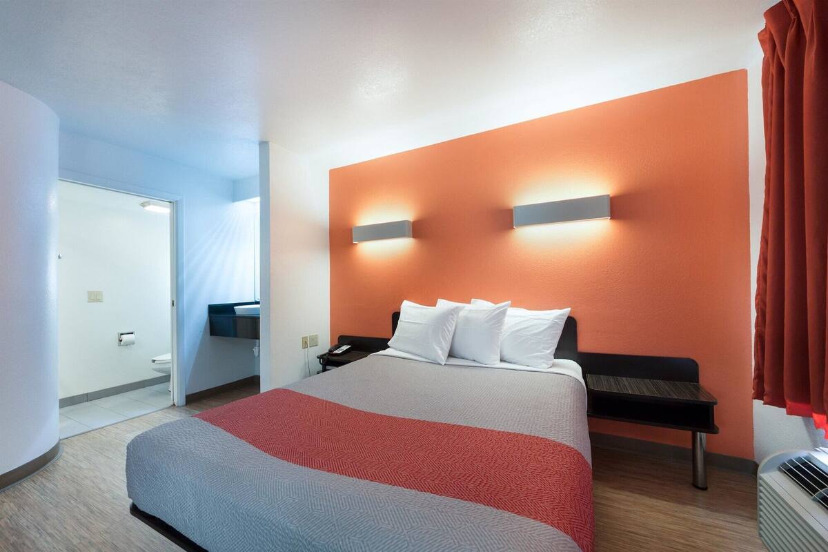 A hotel room with a bed, a nightstand, a wall-mounted lamp, and a window with curtains.