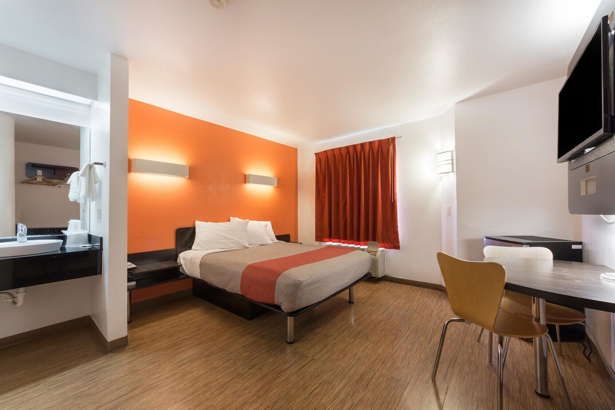 Habitación de hotel con cama, escritorio con silla, televisor, ventana con cortinas y baño con lavamanos y espejo.