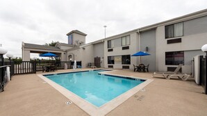 A swimming pool with lounge chairs and umbrellas, surrounded by a building with multiple windows and a fenced area. - Motel 6 Dallas, TX - Northeast (Dallas)
