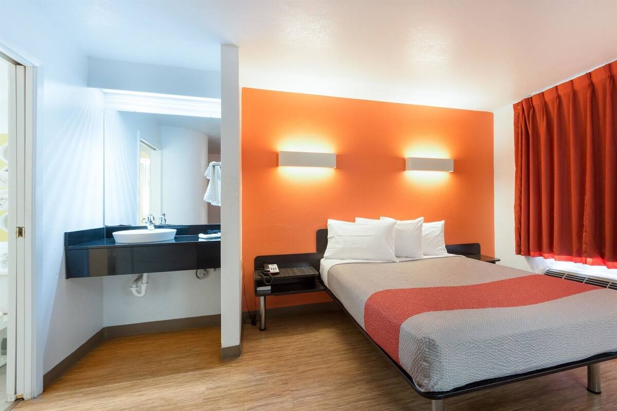 A hotel room with a bed, a nightstand, a bedside table, a bathroom with a sink and mirror, and orange walls.