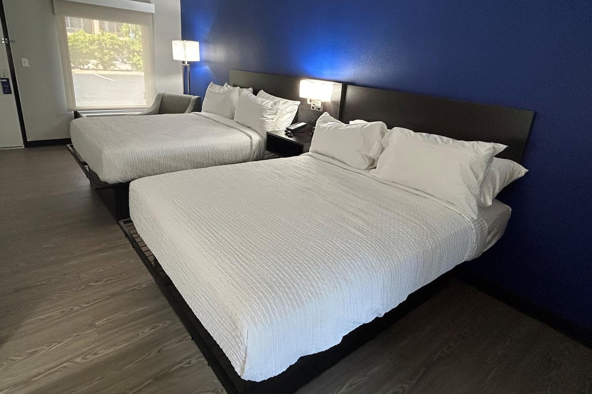 A hotel room with two beds, a nightstand, a lamp, and a view of the outdoors.