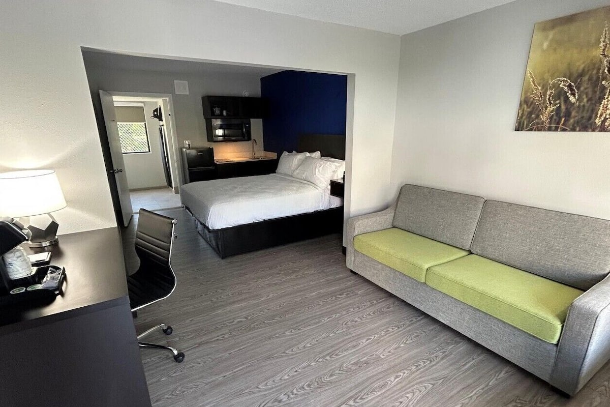 A hotel room with a bed, a grey sofa, a desk with a lamp, and a painting on the wall.