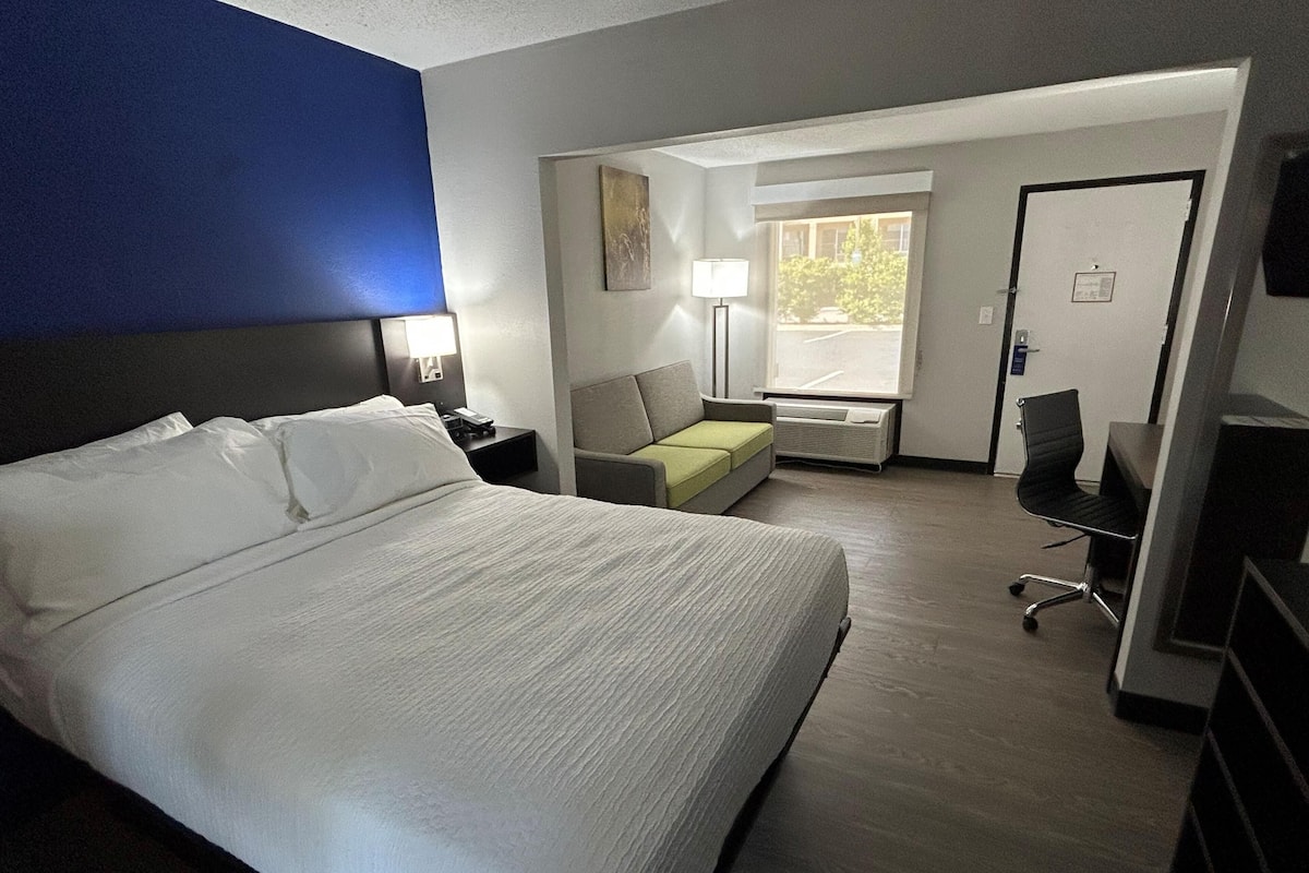 A hotel room with a large bed, a desk, a chair, a sofa, a lamp, and a window.