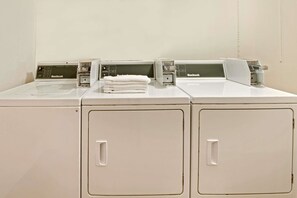 A row of white, front-loading washers and dryers with control panels, stacked towels, and a plain wall. - Super 8 by Wyndham Plano/Dallas Area (Plano)