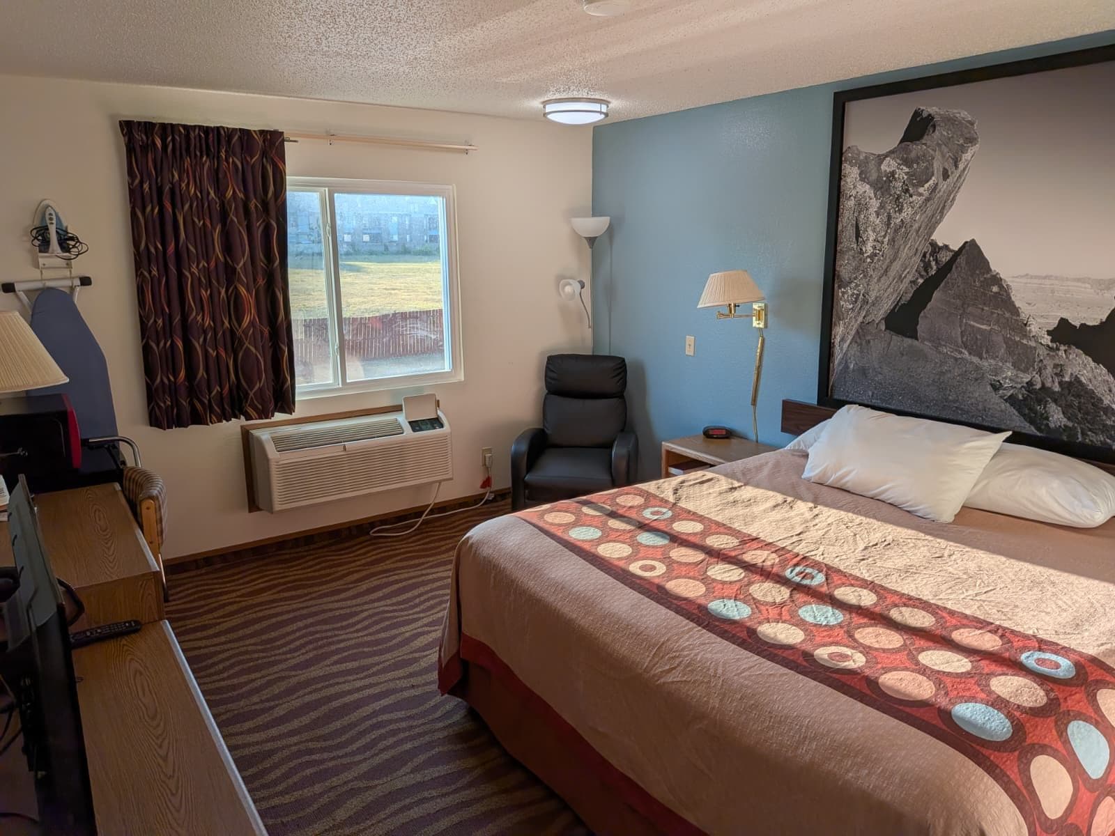 A hotel room with a bed, a desk, a chair, a window with curtains, a wall-mounted lamp, and a large painting.