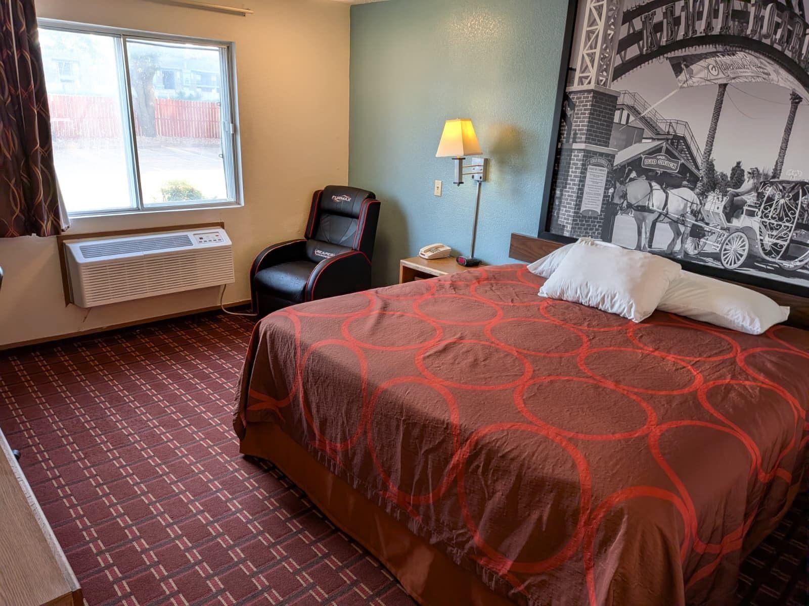 A hotel room with a large bed, a chair, a heater, a lamp, and a wall with a historical photo.
