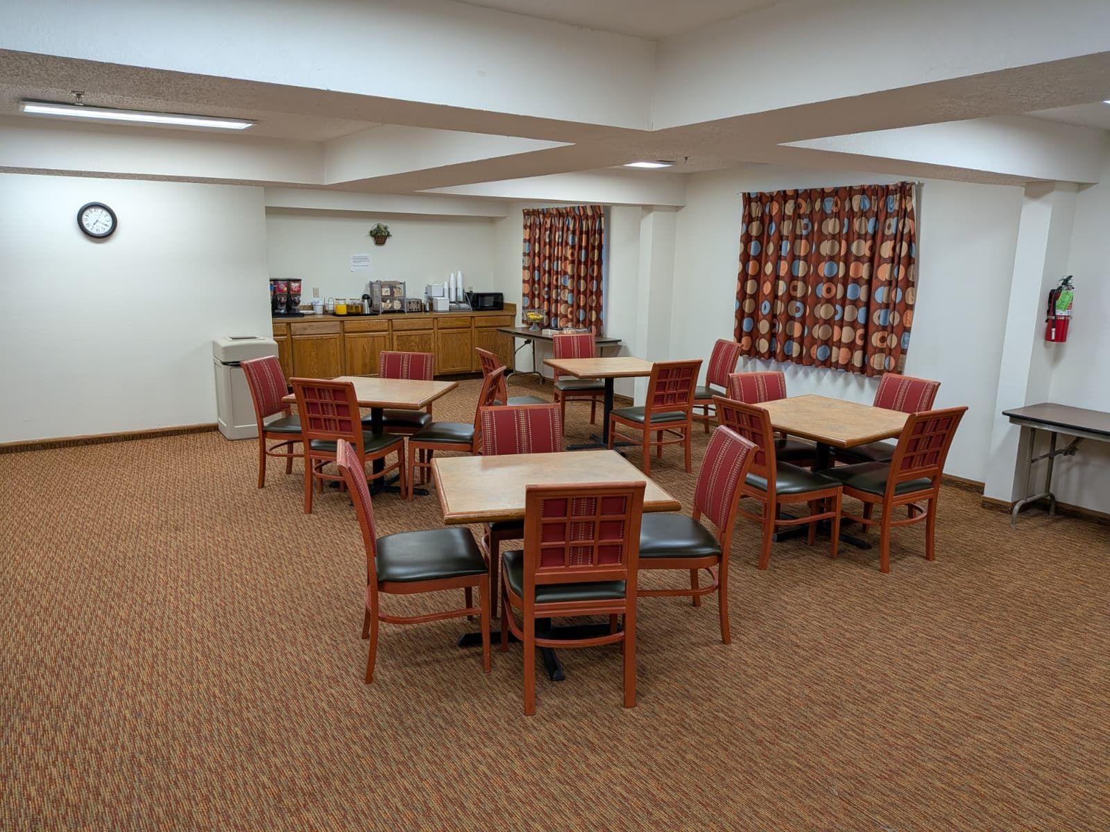 A dining area with multiple tables and chairs, a kitchen in the background, and a fire extinguisher on the wall.