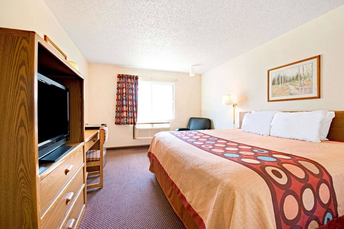 A hotel room with a bed, a desk with a television, a chair, a window with curtains, and a painting on the wall.