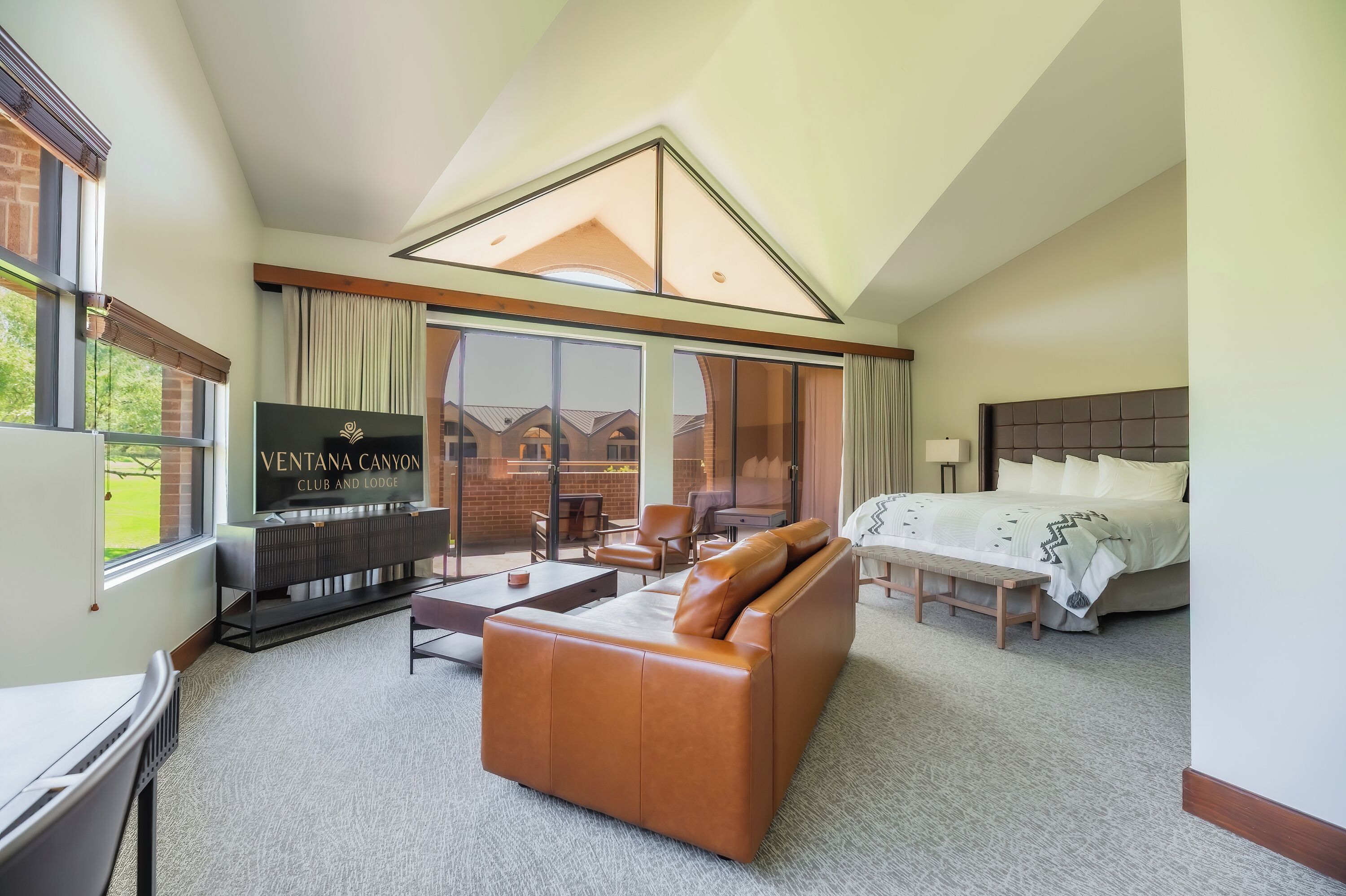 A hotel room with a large bed, a brown leather sofa, a TV, and a balcony with a view of the buildings outside.