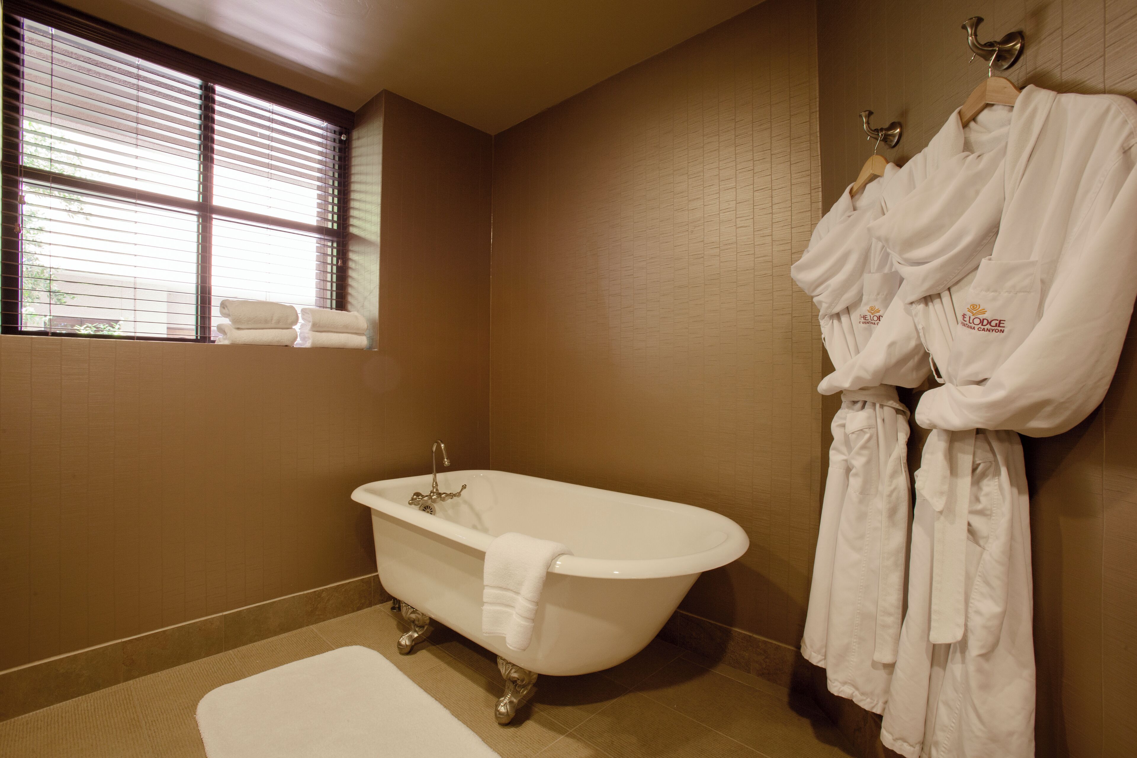 Une salle de bain avec une baignoire sur pieds, une fenêtre avec des stores et un porte-serviettes rempli de peignoirs blancs.
