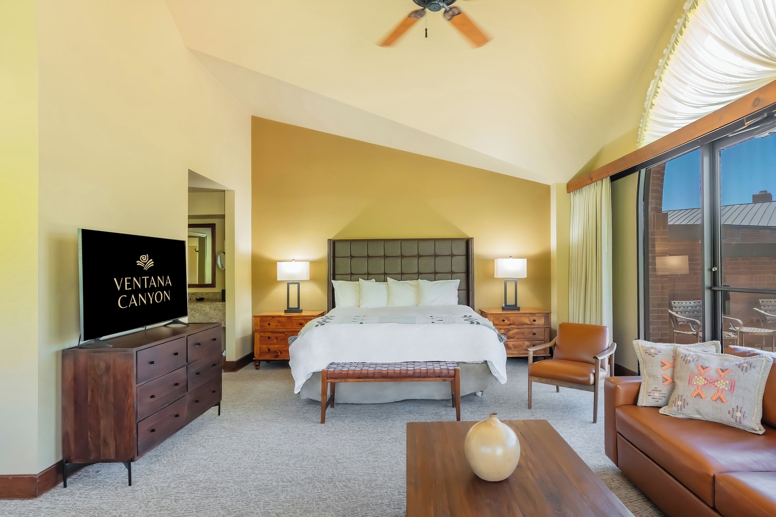 A hotel room with a large bed, a TV, a wooden dresser, a bench, a sofa, and a large window.
