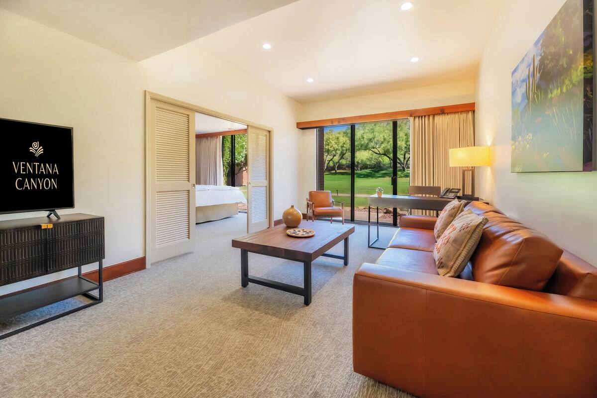 Ein Hotelzimmer mit Fernseher, einer Couch, einem Couchtisch und einem Schreibtisch mit Lampe. Große Fenster bieten Blick auf einen Garten.